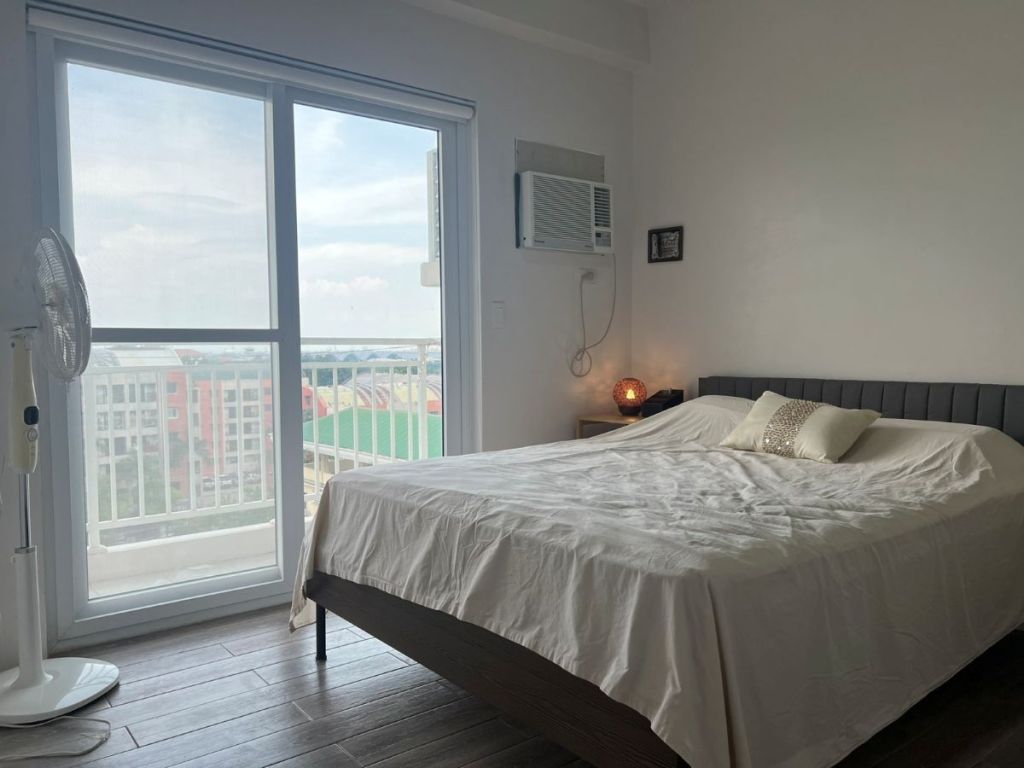 A modern bedroom featuring a neatly made bed with a white blanket and decorative pillow, large windows allowing natural light, a standing fan, and an air conditioning unit on the wall.
