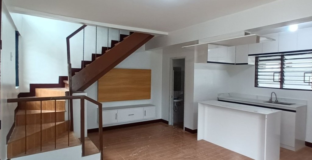 An interior view of a modern kitchen and living area, featuring a staircase, white cabinets, and wooden accents.